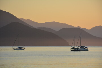 Fototapeta na wymiar dawn with orange landscape, mountain layers and silhouettes of sailboats and boats at sea