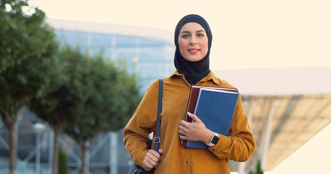 Portrait of young Arabic smiled beautiful woman in black hijab standing at street with textbooks. Pretty muslim female student in traditional Arabian headscarf at city holding books. Outdoor.