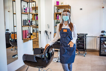 Fototapeta na wymiar Happy small business owner at a hairdressing studio during COVID-19. Portrait of elegant hair salon employee in apron with medical mask, gloves, hair comb and scissors.