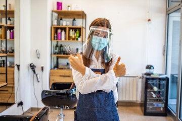 Happy small business owner at a hairdressing studio during COVID-19. Portrait of elegant hair salon employee in apron with medical mask, gloves, hair comb and scissors.