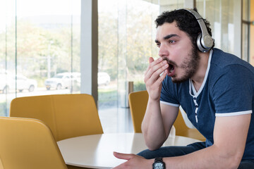 Tired young man yawning with headphones, sitting in the lobby of an office building. Casual young man with a beard.