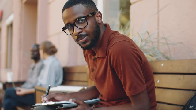 Selective focus shot of young African American businessman in casualwear and glasses taking notes on clipboard and then looking at camera and smiling while working in outdoor cafe