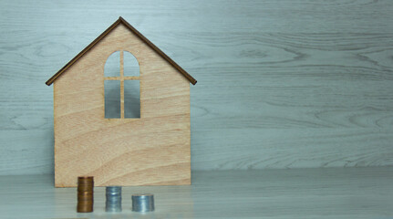 A pile of coins and wooden toy house on a grey background.