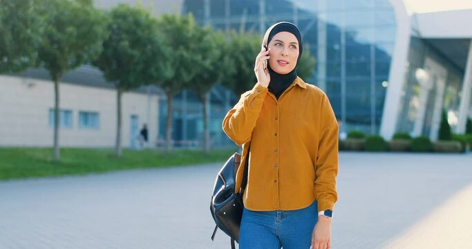 Muslim beautiful young woman in black hijab walking at street and talking on cellpphone. Arabian pretty female strolling the city and speaking on mobile phone outdoors. Telephone conversation.