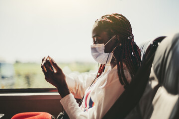 Side view of a young black woman tourist in a virus protective mask traveling in an intercity regular bus on the seat next to the window, she is watching a movie to pass the time, using her smartphone