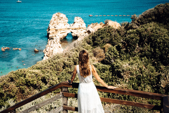 Brown haired girl wearing white dress looking out on the ocean. Lagos, Algarve Coast, Portugal