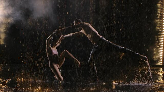 Sexy couple dance contempt with acrobatic movement at dark studio in smoke under spotlight. Silhouettes of wet bodies move sensually among the raindrops.