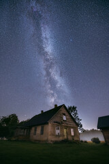 Glowing milky way over crop field covered in fog. Starry night at farmstead. Farm house and tractor.