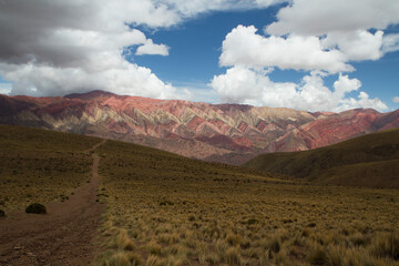 Hiking along the yellow grassland. View of a path across the golden valley leading to the colorful Hornocal mountain under a dramatic cloudy sky. 