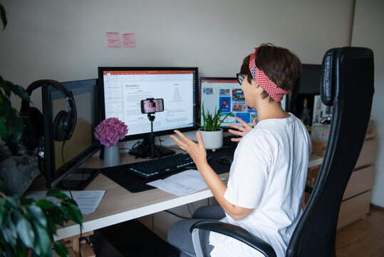 Back view creative blogger shooting video on phone camera sitting at her work desk with few monitors at home working space. E-learning. Remote work concept. Freelancer. Selective focus. Copy space.