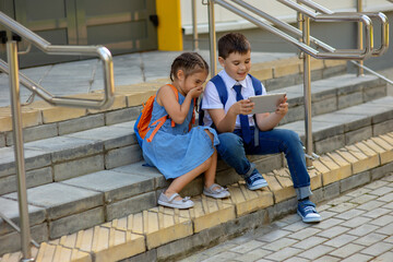 Two schoolchildren a girl with an orange backpack and a boy , laugh, sit on the stairs outside and look into a tablet