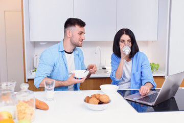 Beautiful happy young couple having breakfast at home and uses laptop at kitchen. Enjoying free time together.