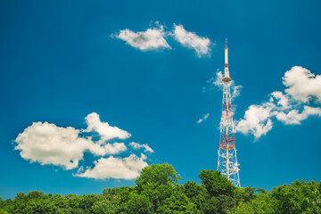 Fototapeta na wymiar TV tower communication infrastructure object urban scenic view clear weather day time with vivid blue sky white clouds and green top of trees bright light