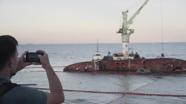 Male blogger is broadcasting live on social networks on journey near sunken tanker Delfi near Odessa. Black Sea coast. Man creating video blog content using smartphone camera about ship accident