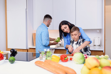 Stylish family making healthy breakfast at kitchen together. Mom teaches son to cut vegetables to salad.