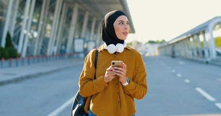 Arabic beautiful woman in hijab and headphones walking at street, tapping on smartphone and messaging. Pretty muslim female in headscarf strolling at aeroport, texting message and chatting. Outdoor. - Powered by Adobe