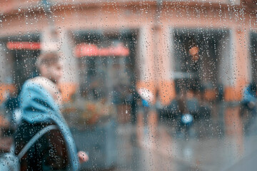 Autumn rainy weather, walking happy young couple together. View through a wet window with raindrops. Concept of modern city, love, lifestyle. Abstract blurred background