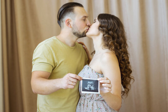 a beautiful curly-haired pregnant girl in a light dress is kissing her husband. They are holding a photo with an ultrasound scan of the future boy.(Selective focus on picture only, people are blurred)