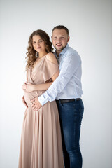 A beautiful curly pregnant girl in an evening dress stands in an embrace with her husband on a white background. She has a long evening dress and he is wearing festive blue shirt. Family Concept