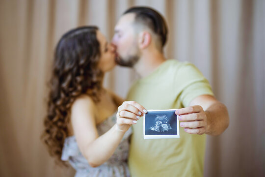 a beautiful curly-haired pregnant girl in a light dress is kissing her husband. They are holding a photo with an ultrasound scan of the future boy.(Selective focus on picture only, people are blurred)