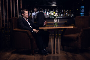 a young man is talking on a smartphone, sitting at a table in a bar alone.