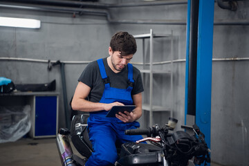 Fototapeta na wymiar A mechanic in work clothes is sitting on a motorcycle. He uses a tablet