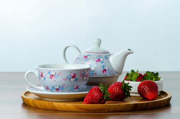 front view of a wooden tray on a table with three strawberries next to a cup of tea with shallow depth of field on a white background