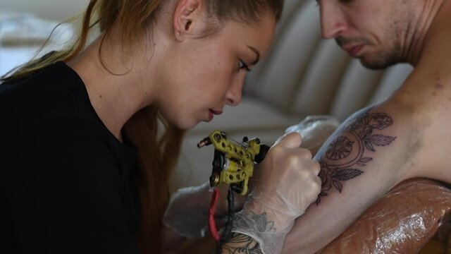 Pretty blonde tattoo artist doing a tattoo to a shirtless man on his shoulder during the day. home tattoo studio.