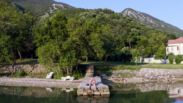 Zoom out shot of a young man lying, getting up on a stone pier in the Kotor bay in Montenegro. Holding a controller and flying a drone shooting the shot. Man by the sea and piloting a drone in 4k.