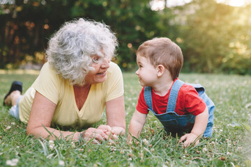 Grandmother lying on grass with grandson boy at home backyard. Bonding of relatives and generation communication. Old woman with baby having fun spending time together outdoors. - 375279351