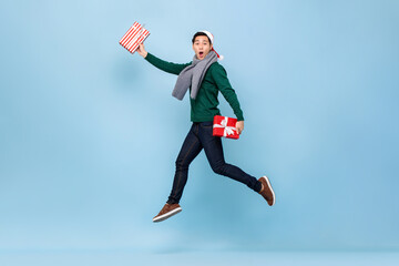 Asian man in Christmas sweater jumping and holding gift boxes isolated in light background