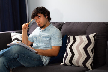 Young hispanic man with wavy hair studying from home writing in a notebook with a pencil, sitting on a sofa