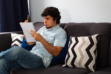 Young hispanic man with wavy hair studying from home writing in a notebook with a pencil, sitting on a sofa