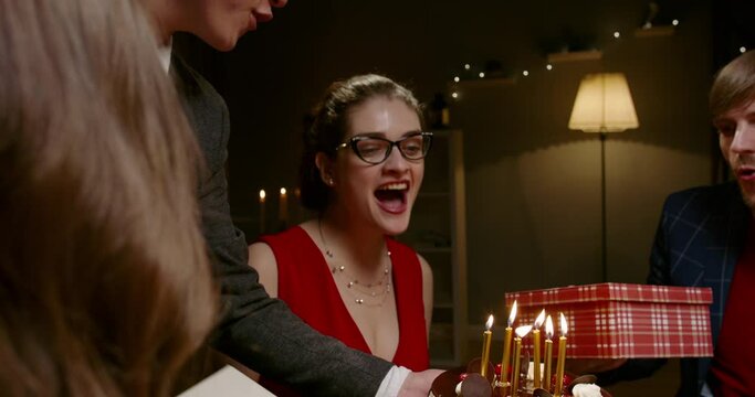 Group of multiethnic friends celebrating birthday. Beautiful caucasian girl is given gifts and blows out candle on special cake - happy people, holidays, friendship concept 4k footage