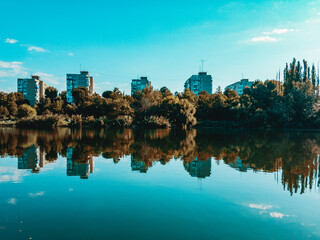Naklejka premium Urban landscape with green vegetation reflected in the water of a reservoir under a cloudless sigim sky.