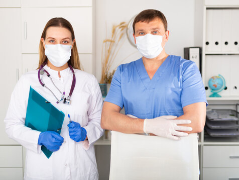 Portrait Of Two Professional Qualified Health Workers Wearing Disposable Surgical Masks And Latex Gloves In Modern Medical Office