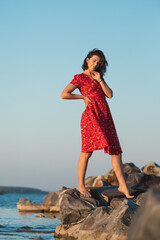 A romantic walk of a curly-haired woman in red dress on a stone against the background of sky and...