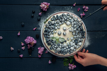 Female hands cut cottage cheese casserole decorated with blackberry, blueberry and acacia flowers on dark wooden table. Flat lay. Copy space.