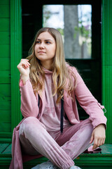 Stylish young girl with blond hair of European appearance sits on the steps, dressed in a pink suit, modern womens fashion, casual clothes