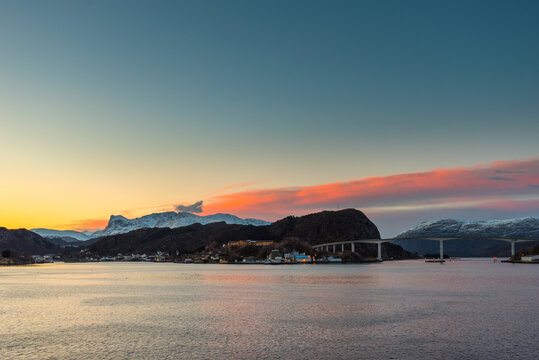 Colorful dawn over the coastline of Norway near the town Maloy with bridge, hills and snow capped mountains seen from cruise ship