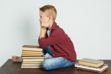 A schoolboy sits on the table near the books, resting his elbows on them and looking away.