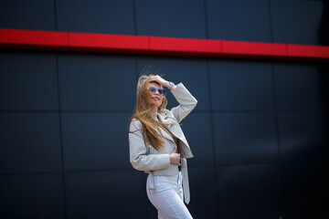 Photo of a long-haired girl in a white blouse and light jeans stands with a smile on the background of the gray wall of the building on a sunny spring day.