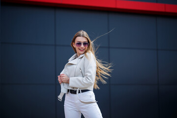 Stylish young woman with long blond hair of European appearance with a smile on her face. Girl in a white jacket and white jeans a warm summer sunny day on a background of a gray building