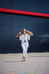 Stylish young woman with long blond hair of European appearance with a smile on her face. Girl in a white jacket and white jeans a warm summer sunny day on a background of a gray building