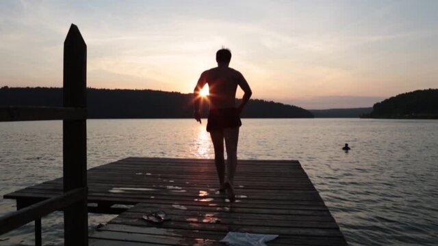 A man jumps from a pier into the sea or lake in the mountains on warm sunny summer day. Slow motion video