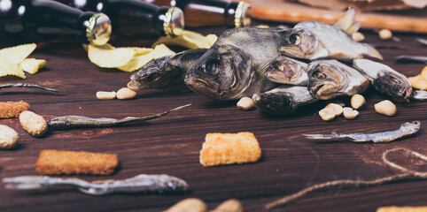 Dried fish, perch, bleak, chips, crackers, nuts and three brown glass bottles of beer on a wooden background.