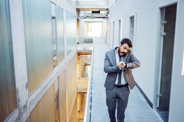 Young bearded businessman looking at wristwatch and talking with business partners. He is late for meeting and making excuse. Building in construction process interior.