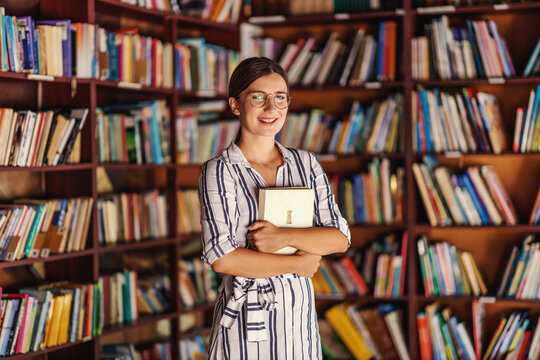 Young attractive smart smiling college girl standing in library and holding book while looking at camera.