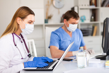 Fototapeta premium Concentrated young female doctor in mask and gloves working on laptop in office, recording diagnosis of patients