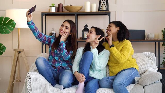 Young diverse women in casual wear sitting on sofa in living room and taking selfie with smartphone, sticking out tongue, pouting lips and showing victory sign. Concept of fun